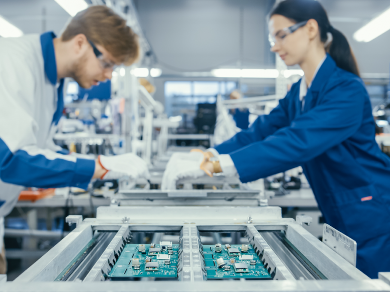 Electronic Assemblers wearing eye protection glasses, discussing as they work at the factory.