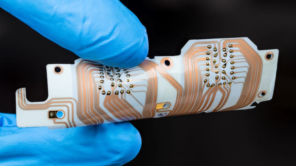 A white flexible Printed Circuit Board (PCB) with an integrated connector is held between the fingers of a technician.